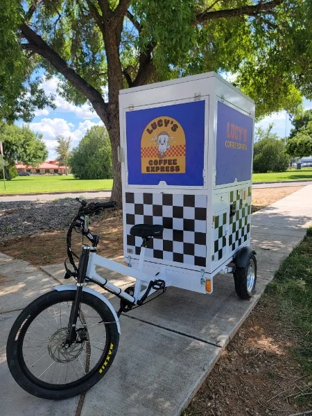 A Lucy's Coffee Express coffee bike parked on a sidewalk under a tree